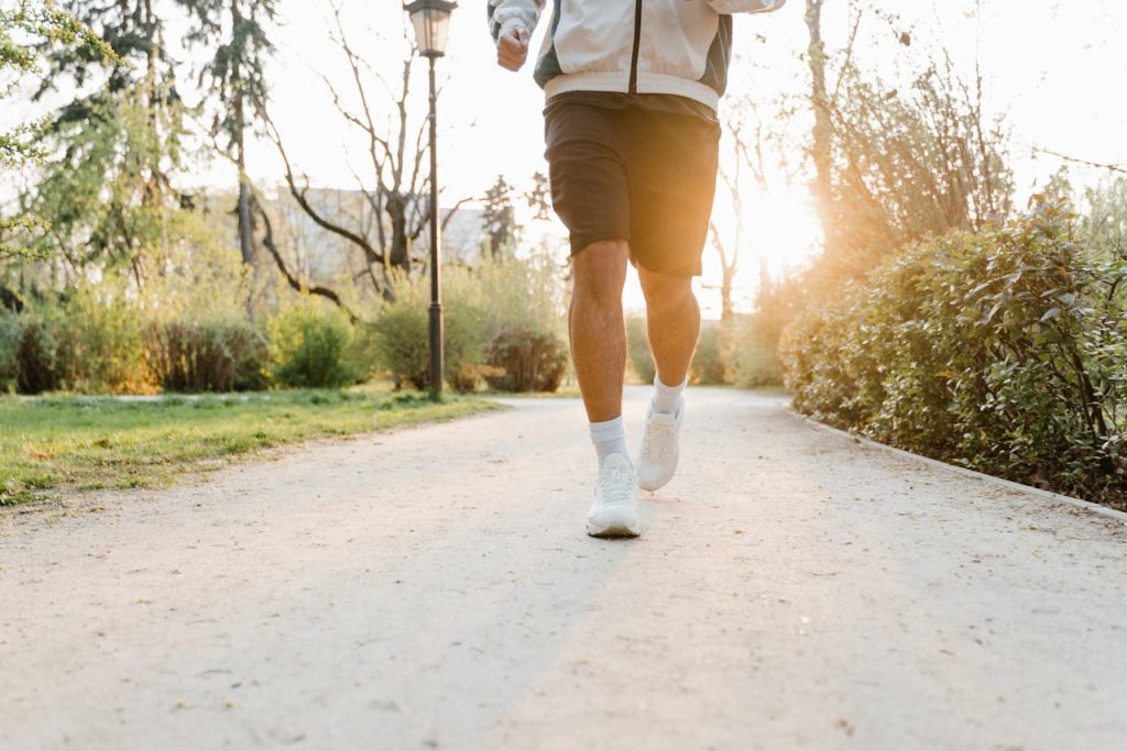 Man jogging on a scenic park trail during sunrise, wearing athletic shorts, a jacket, and white running shoes, embracing an active and healthy lifestyle in nature.
