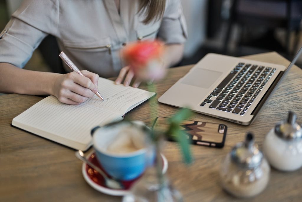 Young woman writing in a notebook while working on a laptop at a cozy café, with a cup of coffee and a smartphone on the wooden table, creating a productive workspace ambiance.