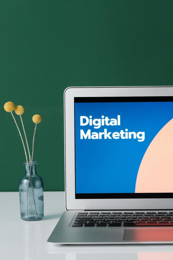 Laptop on a white desk with a digital marketing slide displayed, next to a small glass vase with yellow billy ball flowers and a green backdrop.