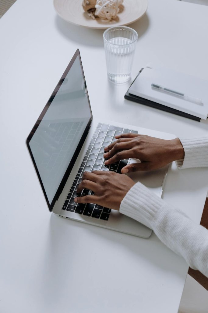 Close-up of hands typing on a laptop at a white desk with a notebook, pen, and glass of water, symbolizing blogging or online writing work.