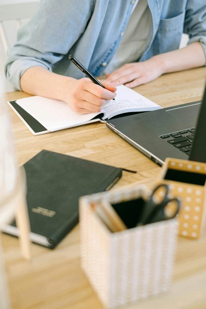 Person writing notes in a notebook at a wooden desk with a laptop, planner, and pen holder, representing blogging or content planning.