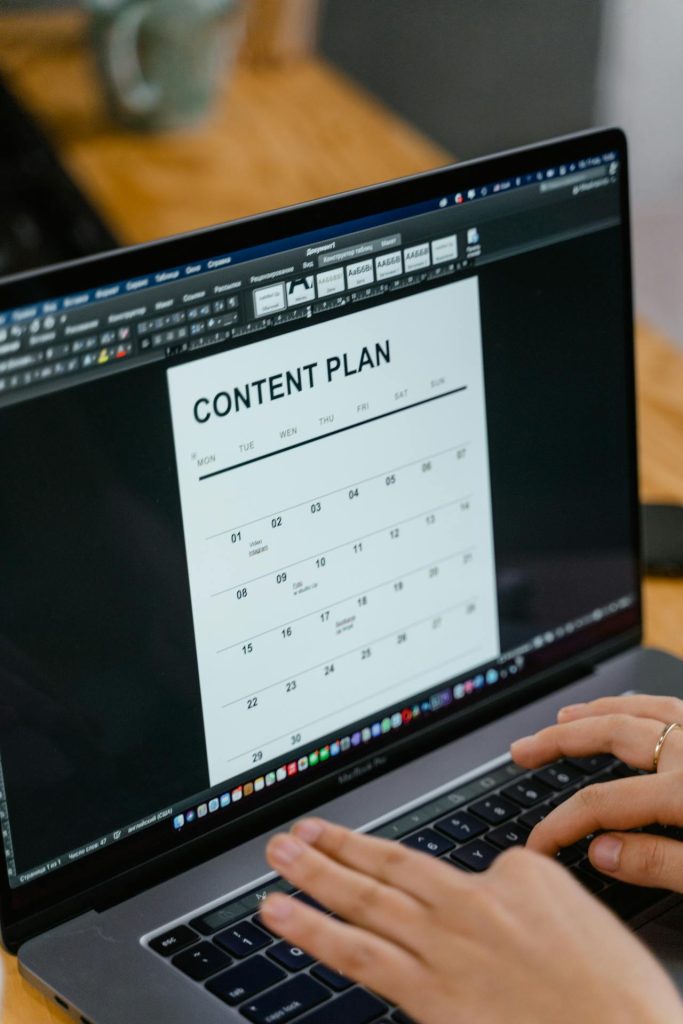 Person typing on a laptop displaying a content calendar layout in a word processing program, working at a wooden desk with a coffee mug nearby.